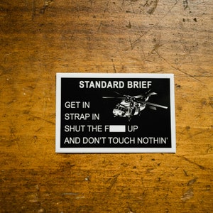 May include: Black and white sticker with a helicopter graphic and the text "STANDARD BRIEF GET IN STRAP IN SHUT THE F UP AND DON'T TOUCH NOTHIN'"