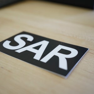 Puede incluir: Una pegatina negra y blanca con la palabra "SAR" impresa en letras blancas.