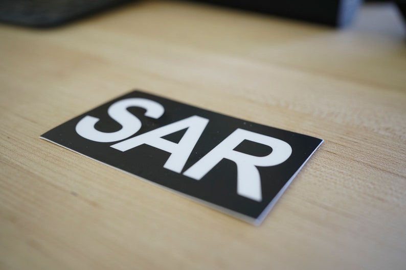 Puede incluir: Una pegatina negra y blanca con la palabra "SAR" impresa en letras blancas.