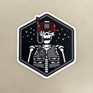 May include: A black and white sticker of a skeleton wearing a life vest and scuba gear. The skeleton has a red and white diving mask and is winking. The background is black with white stars.