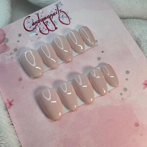 May include: Two sets of oval-shaped, light pink press-on nails are displayed on a pink card. The nails have a glossy finish and are arranged in rows of five. The card has the text "Candymynailz" in a cursive font.