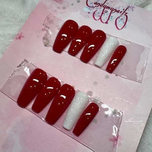 May include: Two sets of press-on nails. Each set features glossy red nails, a glittery white nail, and a red nail. The nails are displayed on a pink card with the text "Candymuniz".