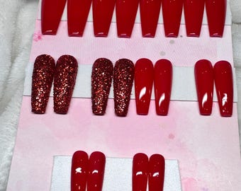 May include: A collection of red press-on nails in various designs. Some nails are solid red, while others are covered in red glitter. The nails are arranged on a pink card with the text "Candymunaiz" in cursive.