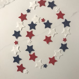 May include: A red, white, and blue felt star garland. The garland is made of felt stars in a spiral pattern. The stars are connected by white string.
