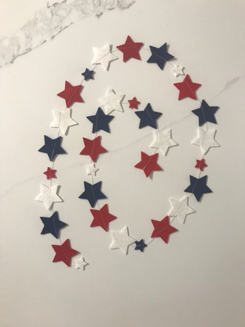 May include: A red, white, and blue felt star garland. The garland is made of felt stars in a spiral pattern. The stars are connected by white string.