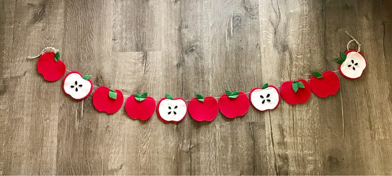 May include: A red felt apple garland with green leaves. The apples are cut in half to show the white inside. The garland is strung on a white twine.