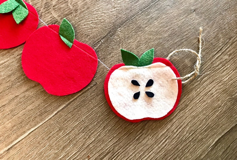May include: A felt apple garland with red apples and a white apple slice with black seeds. The garland is strung with twine.