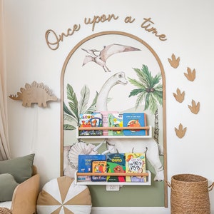 May include: A white bookshelf with two shelves holds a collection of children's books. The bookshelf is mounted on a wall with a green and white dinosaur mural. The mural features a large dinosaur with a long neck and a smaller dinosaur with a long tail. The text "Once upon a time" is written in cursive on the wall above the mural.