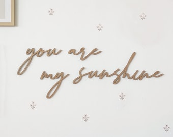May include: Wooden wall decor featuring the words "you are my sunshine" in a cursive script. The background is white with small pink flower decals.
