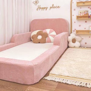 May include: A pink corduroy toddler bed with a headboard and side rails. The bed is decorated with flower-shaped cushions in brown, white, and pink. A cream rug with yellow trim is in front of the bed. The wall has the words "Happy place".