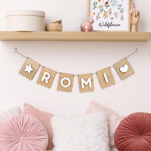 Puede incluir: Bandera de madera con el nombre "ROMI" escrito en letras blancas en banderas individuales. Las banderas están conectadas por una cuerda y tienen bordes festoneados. La primera bandera tiene una estrella blanca y la última bandera tiene un corazón blanco.