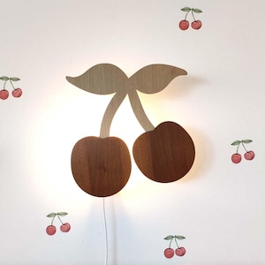 Puede incluir: Una lámpara de pared de madera con forma de cerezas con un acabado marrón claro. La lámpara tiene forma de dos cerezas con hojas verdes. Hay varias pegatinas pequeñas de cerezas rojas en la pared detrás de la lámpara.
