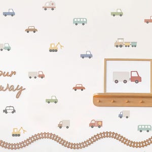 May include: A wall adorned with pastel-coloured vehicle decals, such as cars, trucks, buses, and an ambulance. A framed picture of a truck sits on a wooden shelf. A wooden train track and the words "our way" are also on the wall.