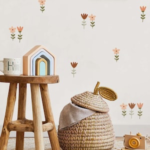 Puede incluir: Un taburete de madera sostiene una taza con la letra "H" y un conjunto de arcos de madera en colores pastel. También se ven una cesta tejida con tapa y una cámara de juguete de madera. El fondo es una pared blanca decorada con calcomanías florales.
