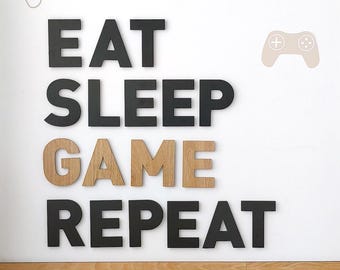 May include: Black and brown wooden letters spelling out "Eat Sleep Game Repeat" on a white wall with a black and white game controller graphic.