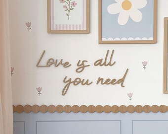 May include: Wooden wall decor featuring the phrase "Love is all you need" in cursive script. The decor is mounted on a white wall with a light blue trim and a scalloped wooden border. There are small pink flower illustrations on the wall.