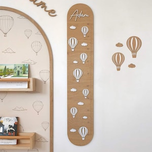 Pode incluir: Gráfico de altura de madeira com acabamento castanho claro, com balões de ar quente e nuvens brancas. O gráfico é personalizado com o nome "Adam" no topo e inclui medidas em centímetros. Arte de parede decorativa para o quarto de uma criança.