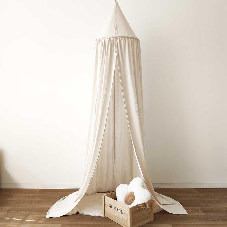 May include: A white cotton canopy bed curtain with a wooden storage box and a white plush flower-shaped pillow inside. The canopy is hanging from the ceiling and is draped over the bed.