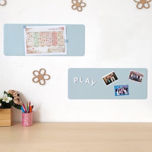 Peut inclure: Deux tableaux mémo rectangulaires bleu clair sont montés sur un mur blanc. L'un contient un document imprimé, fixé avec une pince bleue. L'autre porte le mot "PLAY" en lettres blanches et trois photos.