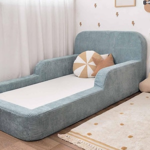 Op de afbeelding: Een lichtblauw peuterbed van corduroy met een hoofdbord en zijrails. Het bed heeft een witte matras en is versierd met een rond, gestreept kussen en een stervormig kussen. Het bed staat op een crèmekleurig tapijt.