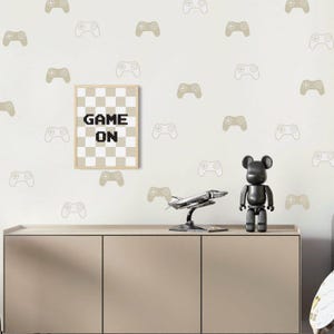 Könnte beinhalten: Ein beiger Schrank mit einem gerahmten Druck mit der Aufschrift "GAME ON" in schwarzen Pixelbuchstaben. Der Druck hat einen karierten Hintergrund. Die Wand ist mit Aufklebern von Videospiel-Controllern in Beige und Weiß dekoriert. Eine schwarze Bärenfigur und ein silbernes Modellflugzeug stehen auf dem Schrank.