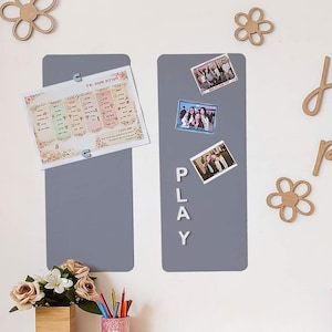 Peut inclure: Deux tableaux magnétiques gris avec des photos et le mot "PLAY" en lettres blanches. Un tableau contient une feuille imprimée avec du texte hébreu. Des découpes de fleurs et de lettres en bois décorent le mur blanc.