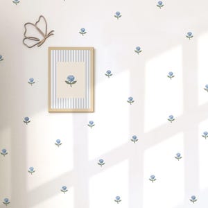 May include: A framed picture with a blue flower illustration hangs on a white wall adorned with blue flower decals. A wooden butterfly decoration is also on the wall. The picture frame has a light wood finish and a striped background.