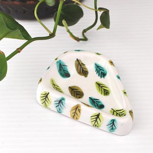 May include: A triangular ceramic dish with a white base featuring a repeating pattern of small, hand-painted leaves in shades of teal, olive green, and lime green. The dish has a smooth, glossy finish and slightly raised edges, forming a shallow bowl.