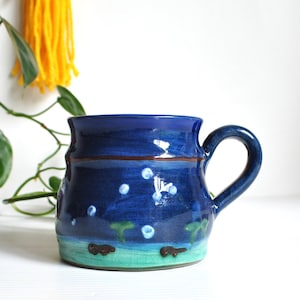 May include: A ceramic mug with a glossy deep blue glaze featuring a hand-painted night sky motif. The design includes small white dots representing stars, a thin brown horizontal line, and a light green base with dark brown animal silhouettes.