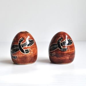 May include: Two brown, egg-shaped ceramic salt and pepper shakers. Each shaker features a black lizard design. The shakers have a glossy finish and are set against a white background. The lizard design is a stylized, tribal-inspired illustration.