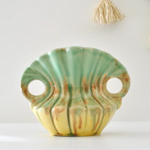 May include: A ceramic vase shaped like a scalloped shell with two circular side handles. The glaze features a gradient transition from a pale yellow base to a soft green top, accented with vertical streaks of brown throughout the ribbed surface.