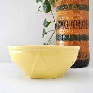 May include: A pale yellow ceramic bowl with a faceted geometric exterior design. The bowl features a smooth, glossy finish and a wide, circular rim, positioned in front of a tall, cylindrical brown and orange textured ceramic vase.