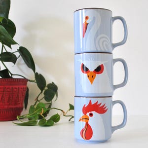 May include: A vertical stack of three light blue ceramic mugs, each featuring a different animal illustration. The top mug displays a white swan, the middle mug shows a white owl with red eyes, and the bottom mug depicts a white rooster with a red comb.