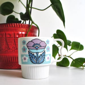 May include: A white ceramic mug featuring a stylised floral graphic in shades of purple, light blue, and dark blue. The mug has a cylindrical body with a ribbed base and a curved handle. The floral design is centred on the front of the mug.