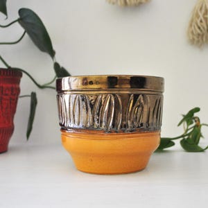 May include: A ceramic planter featuring a two-tone design with a bright orange base and a metallic bronze upper section. The upper portion is decorated with a repeating vertical carved leaf pattern that encircles the entire circumference of the pot.