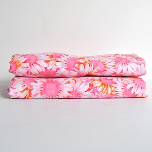 May include: A folded piece of fabric featuring a vibrant pattern of pink and white daisy-like flowers with orange accents. The textile is neatly stacked in two layers, displaying a repeating floral motif across the entire visible surface area.