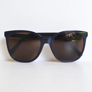 May include: Navy blue sunglasses with brown lenses. The sunglasses have a classic, slightly oversized frame design. The lenses are a dark brown colour, providing a contrast to the navy blue frame. The sunglasses are set against a white background.