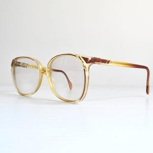 May include: Vintage spectacles with a clear, oversized frame in a light yellow colour. The temples and upper frame have a brown accent. The lenses are clear, and the overall design is retro.