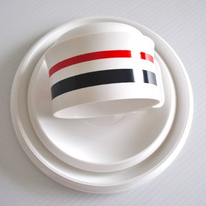 May include: White ceramic lid with three red, black, and white stripes. The lid is sitting on a white saucer.