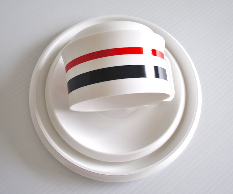 May include: White ceramic lid with three red, black, and white stripes. The lid is sitting on a white saucer.