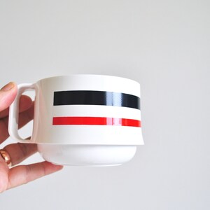 May include: White ceramic mug with a black and red stripe design. The mug has a handle and a wide base.