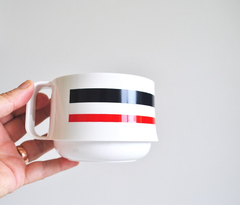 May include: White ceramic mug with a black and red stripe design. The mug has a handle and a wide base.