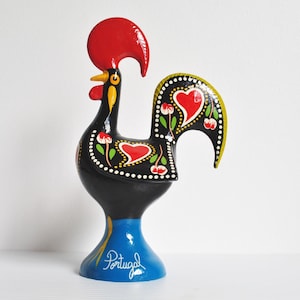 May include: A ceramic rooster ornament. The rooster is painted black with a red comb and yellow, white, and green details. The tail has a heart design. The base is blue and inscribed with the word "Portugal".