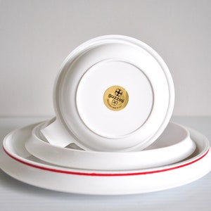 May include: A white ceramic cup and saucer set with a red rim. The saucer has a gold sticker with the text "guzzini (IT)" and a logo.