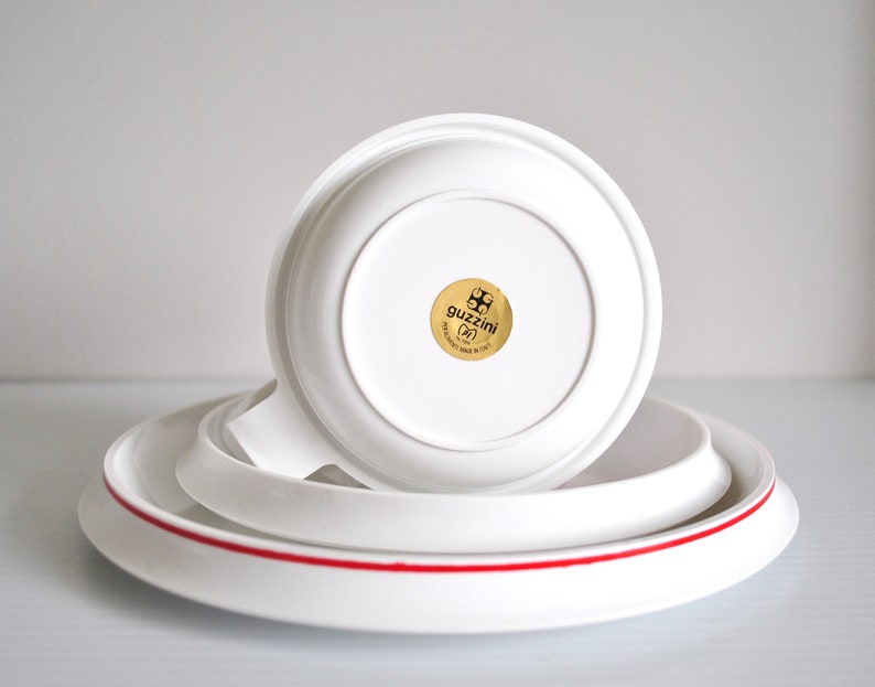 May include: A white ceramic cup and saucer set with a red rim. The saucer has a gold sticker with the text "guzzini (IT)" and a logo.