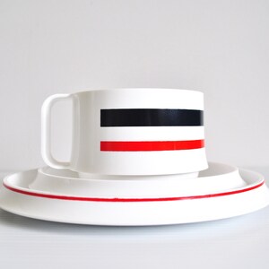 May include: White ceramic teacup and saucer set with black and red stripes. The cup has a handle and the saucer has a red stripe around the edge.