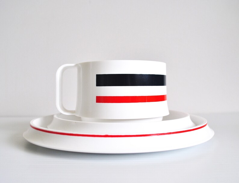 May include: White ceramic teacup and saucer set with black and red stripes. The cup has a handle and the saucer has a red stripe around the edge.