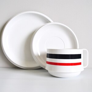 May include: A white ceramic cup with a black and red stripe design sits on a white saucer. The saucer is nested inside a larger white saucer.