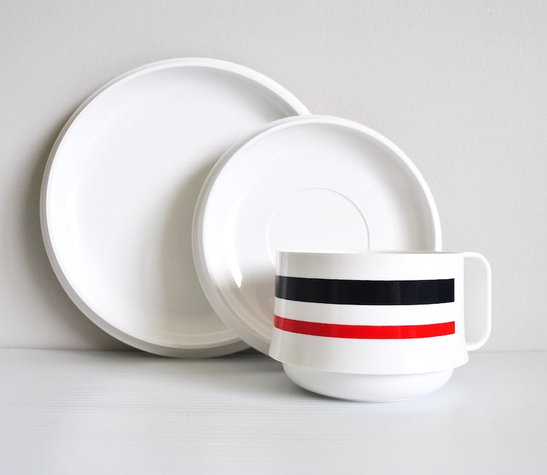 May include: A white ceramic cup with a black and red stripe design sits on a white saucer. The saucer is nested inside a larger white saucer.