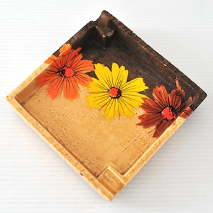May include: A square ceramic ashtray featuring a gradient finish that transitions from a light tan base to a dark brown rim. The interior surface is decorated with three painted flowers in vibrant shades of orange, yellow, and deep red.